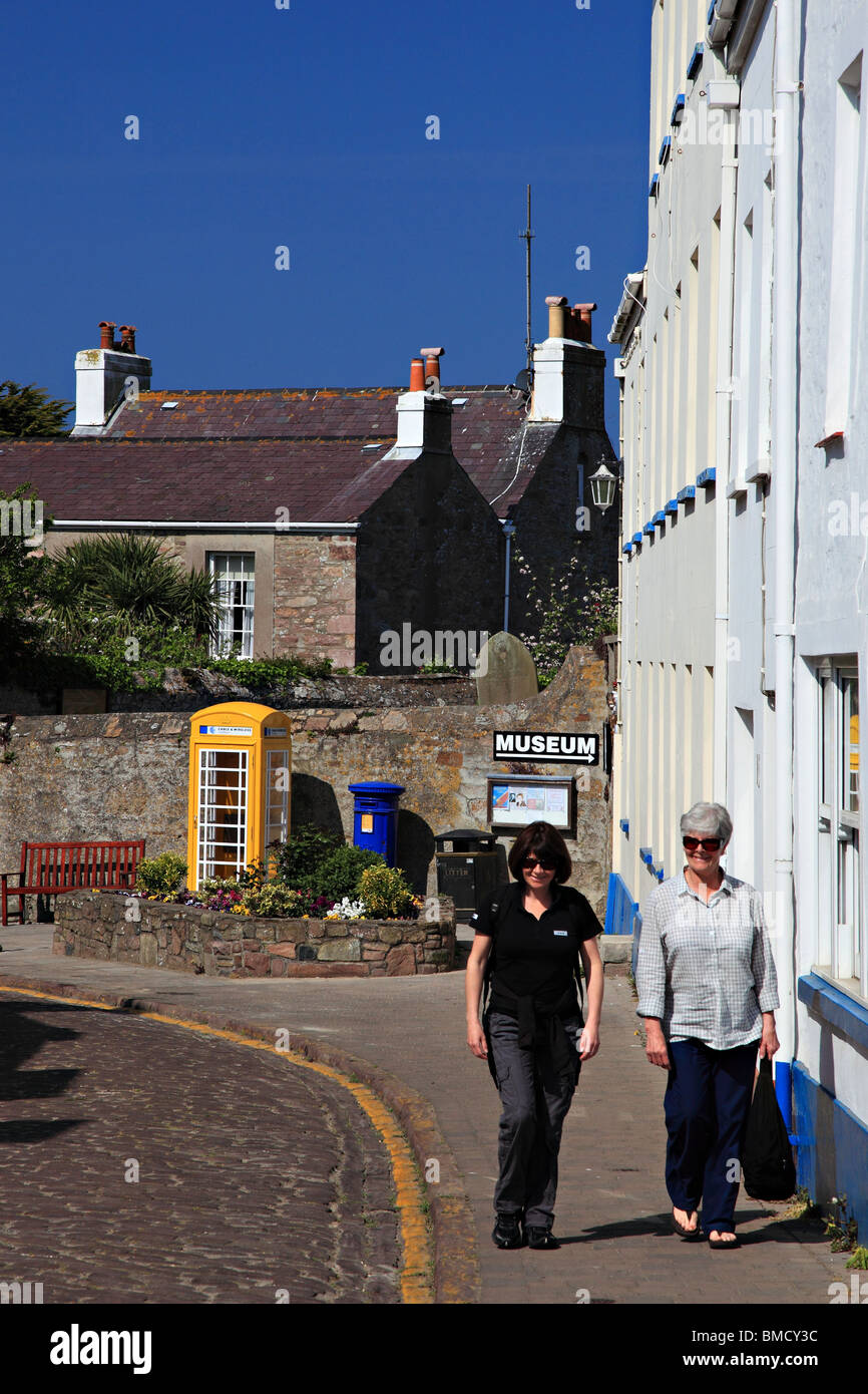 Frauen gehen auf High Street von St. Anna mit erhltlich Telefon Box und blau Brief Kasten, Alderney, Kanalinseln, Großbritannien Stockfoto