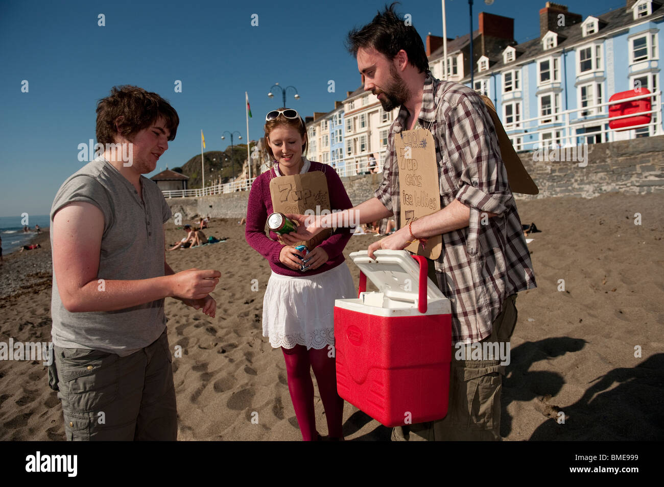 zwei unternehmungslustige junge Studenten, kalte Getränke und Eis am Stiehl an Menschen am Strand von Aberystwyth, auf einen heißen Sommer Nachmittag UK zu verkaufen Stockfoto