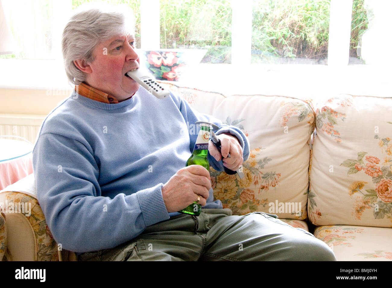 Reifer Mann TV Fernbedienung in den Mund zu halten, während eine Flasche Bier zu öffnen Stockfoto