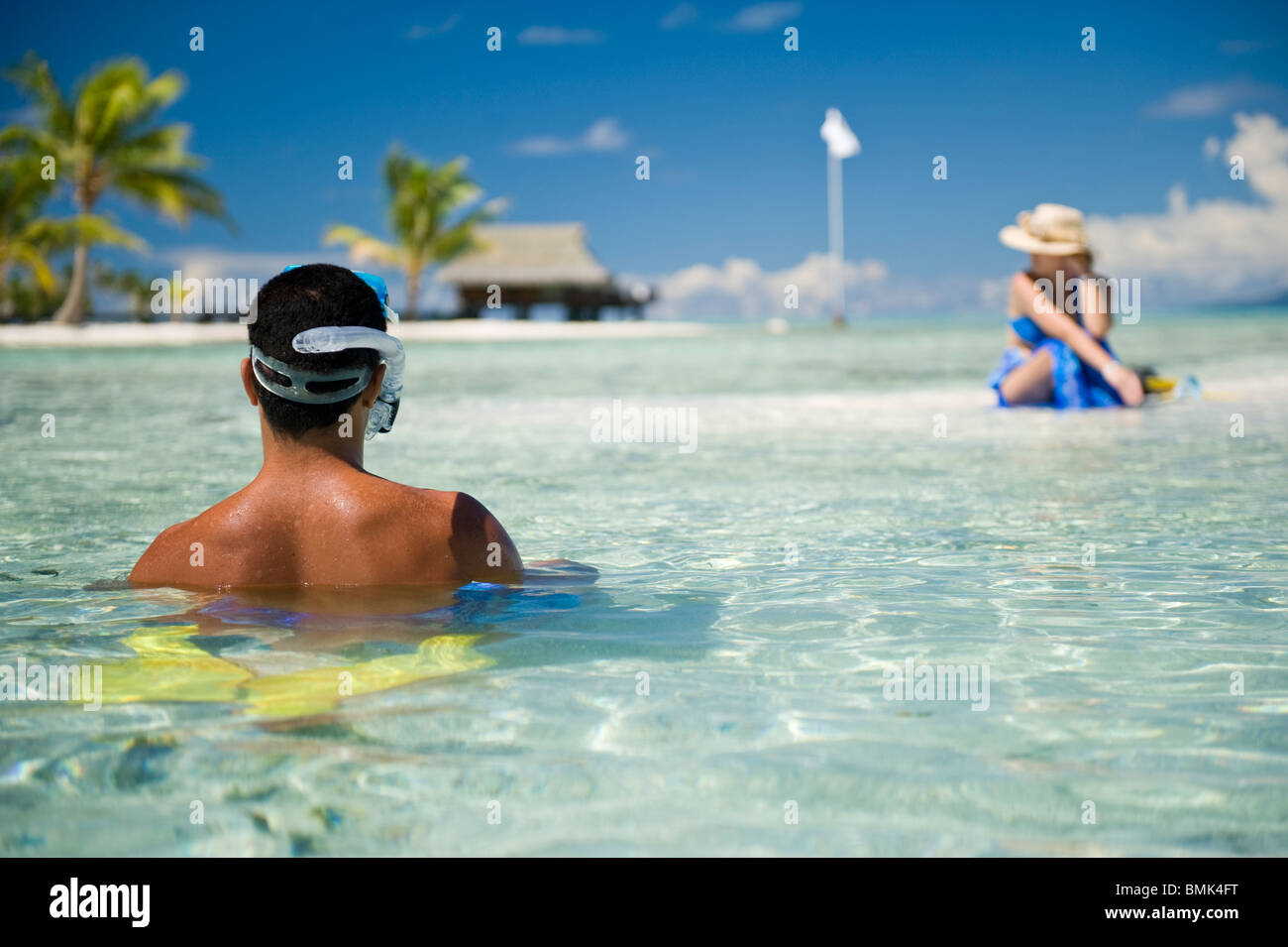ein Mann und eine Frau sitzen im seichten Wasser an einem weißen Sandstrand. Die Frau ist Sonnenbaden, der Mann geht zum Schnorcheln Stockfoto