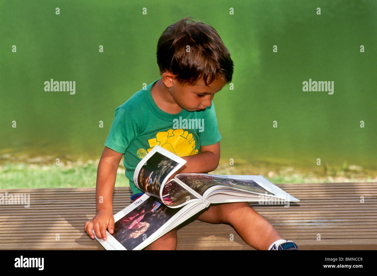 Kleiner Junge sitzen draußen auf einer Terrasse in der Nähe von See Blick mit Interesse auf ein Bilderbuch, ruhig lernen, Midwest USA Stockfoto