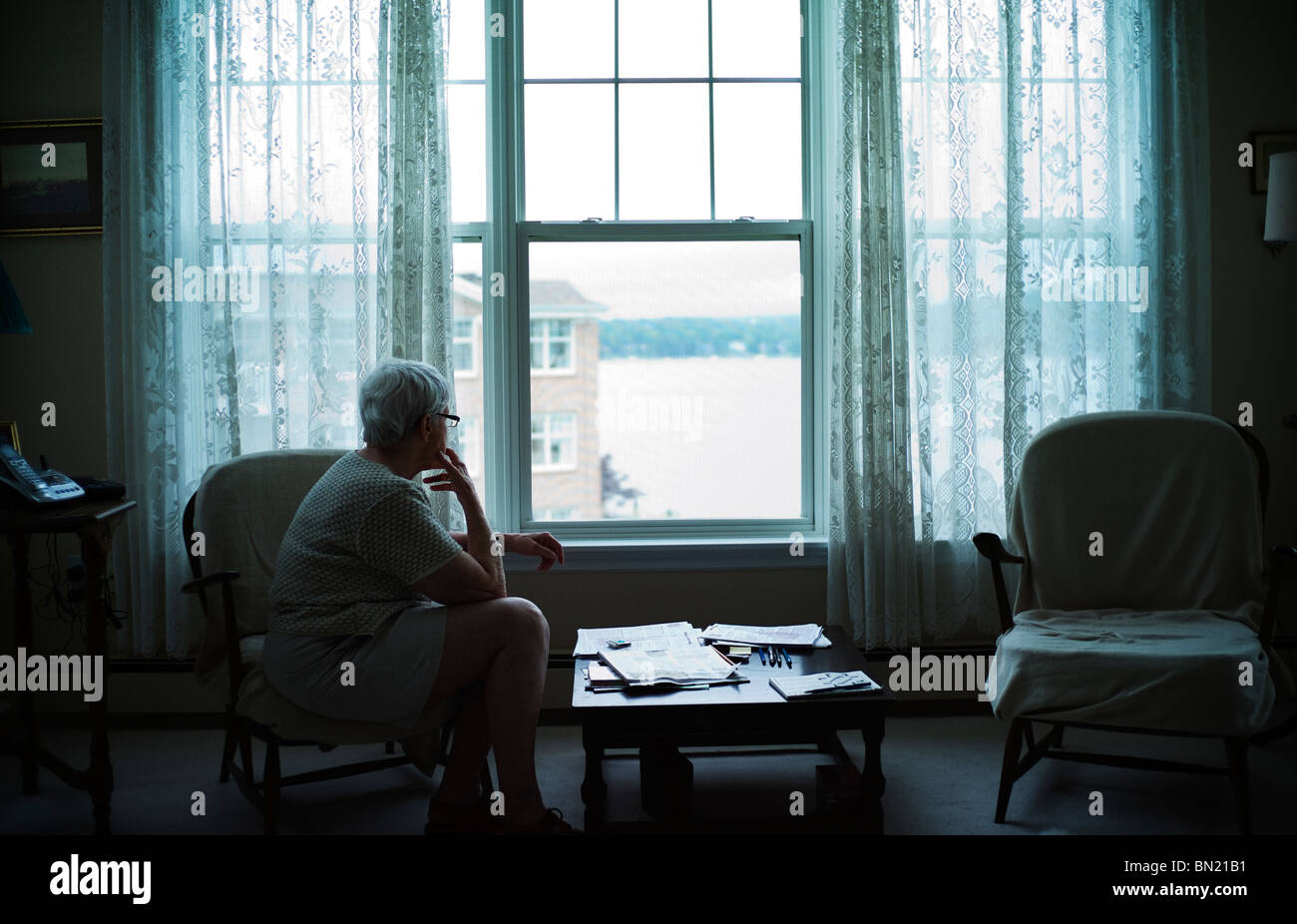 Alte Frau sitzt in schwach beleuchteten Wohnzimmer, starrte aus dem Fenster Stockfoto
