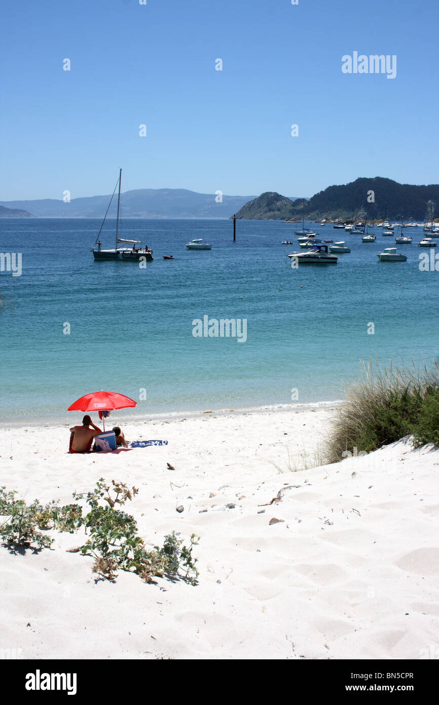 Strand Praia de Rodas, mit paar unter roten Sonnenschirm und Boote in der Bucht von Vigo, Galicien, Nordspanien, Illa de Faro, Illas Cies Stockfoto