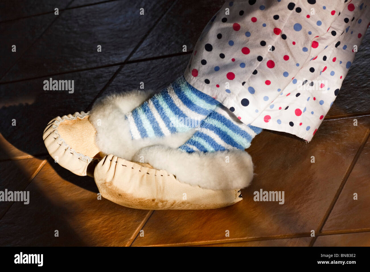 Eine Frau trägt Pelz gefütterten Mokassin Hausschuhe, gestreifte Candy Socken und gepunkteten Schlafanzug mit ihren Füßen in der Sonne. Stockfoto