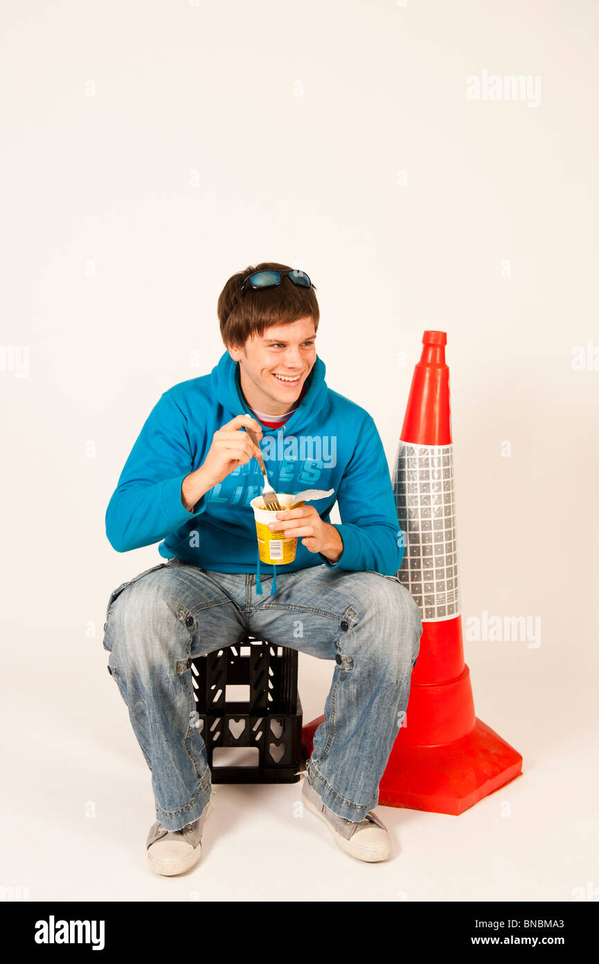 Junger Mann Essen Topf Nudeln warme Snacks sitzen auf einer Kunststoff Kiste neben eine orange Verkehr Kegel, UK Stockfoto
