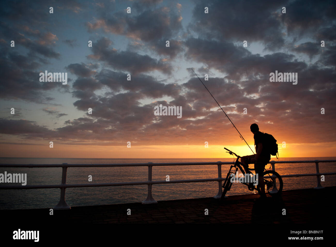 Die Silhouette eines jungen Mannes mit dem Fahrrad bei Sonnenuntergang, etwa zum Angeln, Cardigan Bay West Wales UK Stockfoto