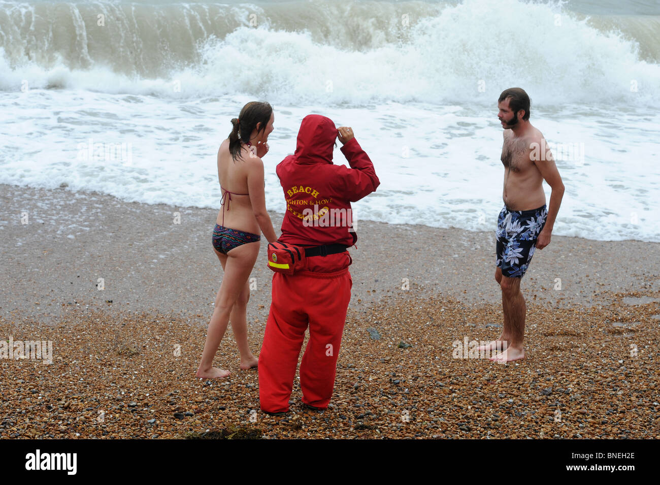 Junge Frau im Bikini und Mann, zwanziger im Gespräch mit Rettungsschwimmer am Strand von Brighton, vor dem Schlafengehen in der rauen See. Stockfoto