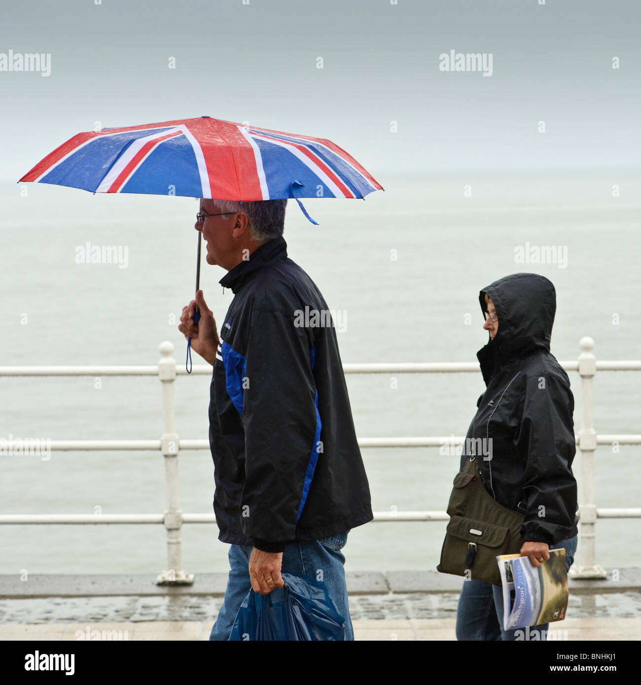 Menschen zu Fuß entlang Aberystwyth promenade im Regen eine Juli nass Sommernachmittag, Wales UK Union Jack Dach festhalten Stockfoto