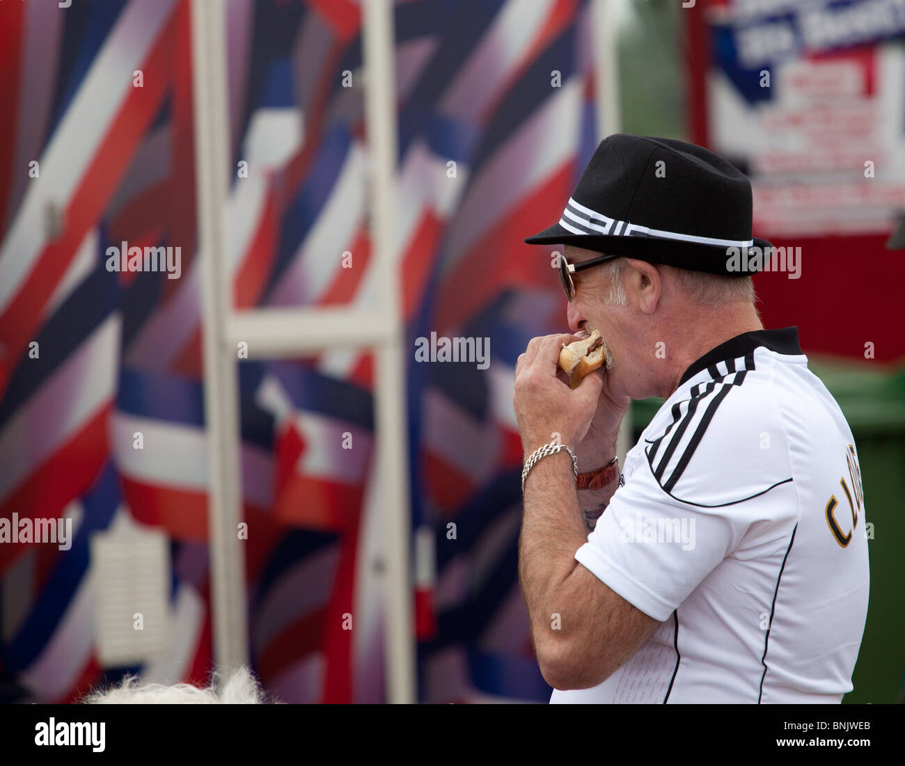 Ein Mann, der ein Fußballtrikot trägt, nimmt einen großen Bissen von EINEM Burger, während er bei einer Open-Air-Veranstaltung in Fairford, großbritannien, aus einem Fast-Food-Outlet geht Stockfoto