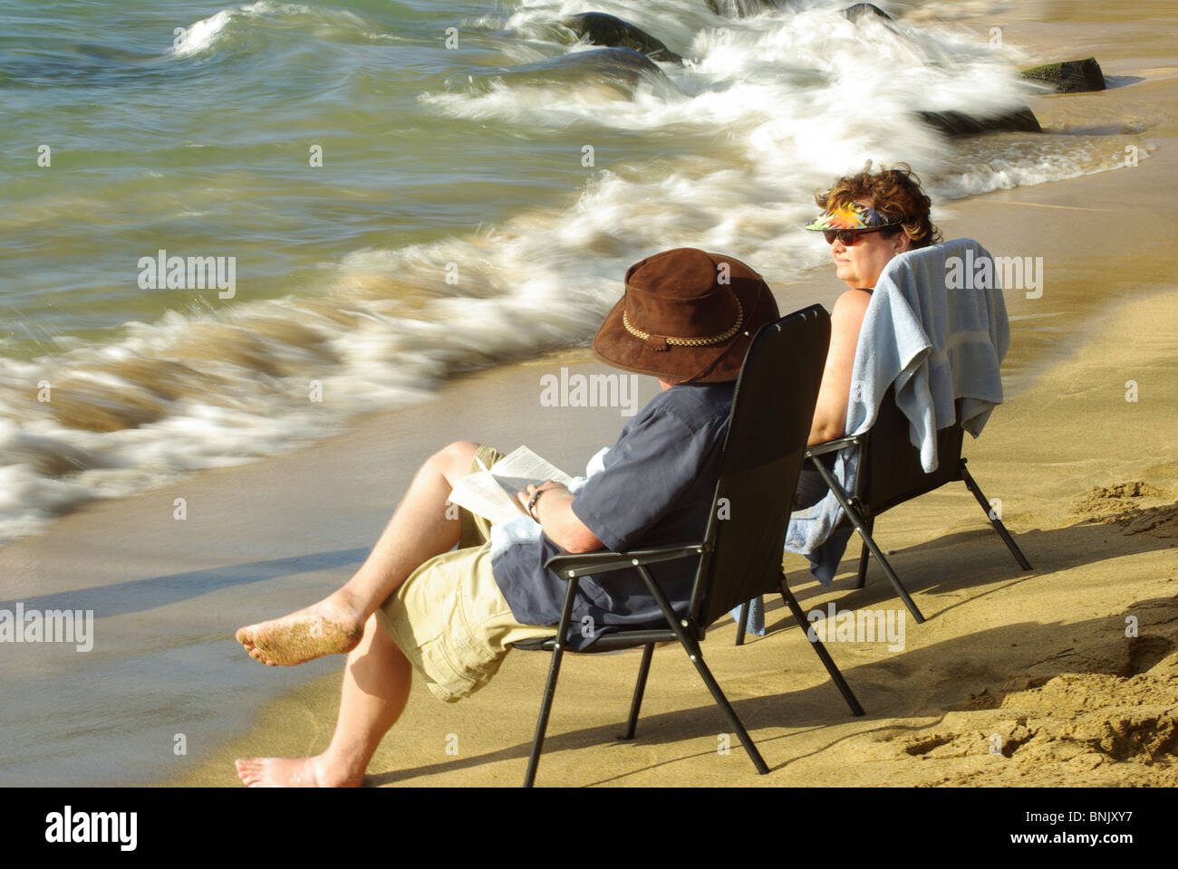 mittleren gealterten paar sitzen auf Stühlen am Strand entspannen, einer Lesung, winkt ein Blick auf den Ozean stürzt auf den Strand. Stockfoto