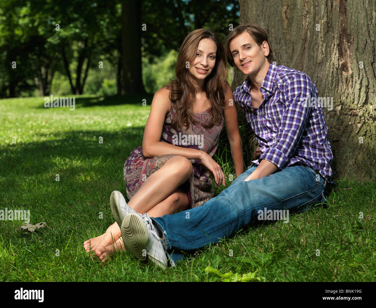 Junge glückliche Paar in ihren frühen dreißiger Jahren sitzen auf Rasen unter einem Baum in einem park Stockfoto