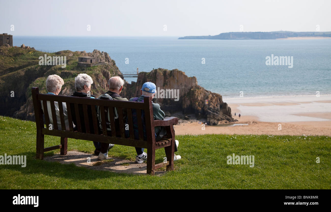 Vier ältere Menschen sitzen in der Sonne auf der Bank am Meer Tenby Wales UK Stockfoto