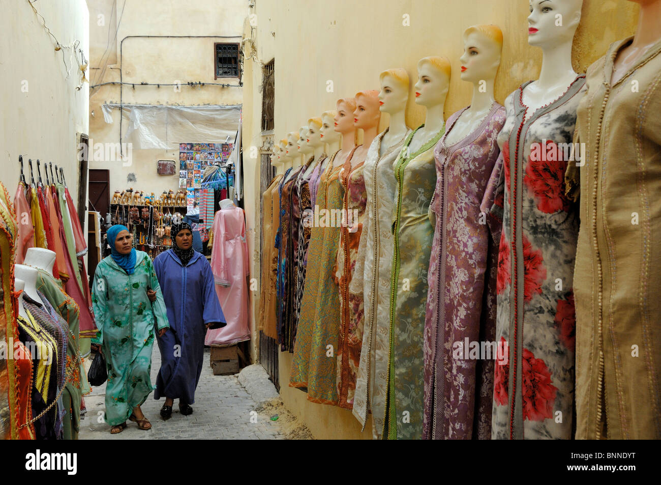 Zwei marokkanische Frauen, die an Mode- oder Bekleidungsgeschäft vorbeigehen, verkaufen traditionelle ethnische Kleidung oder ethnische Kleider, Souk, Medina, Fez, Marokko Stockfoto