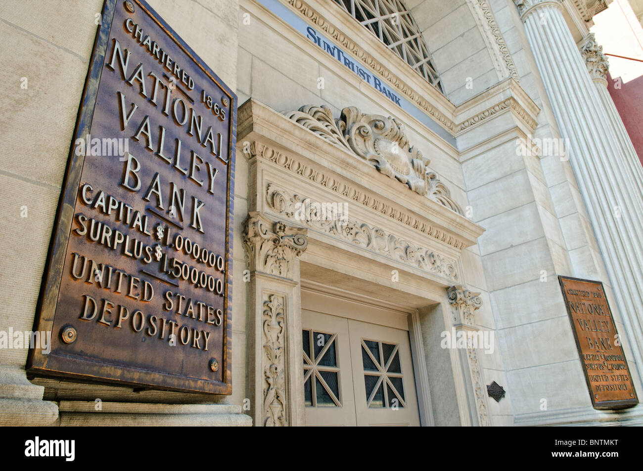 National Valley Bank Sign Staunton // STAUNTON, Virginia — das historische Bronzeschild der National Valley Bank, das 1865 gechartert wurde, ist an seinem Gebäude in der Innenstadt von Staunton zu sehen. Das Zeichen wirbt stolz auf das Kapital der Bank von 1.000.000 Dollar und einen Überschuss von 1.500.000 Dollar, was ihre bedeutende finanzielle Stellung widerspiegelt. Die kunstvoll verzierte klassische Architektur des Gebäudes, heute Sitz der SunTrust Bank, trägt zu seiner historischen Präsenz bei. Stockfoto