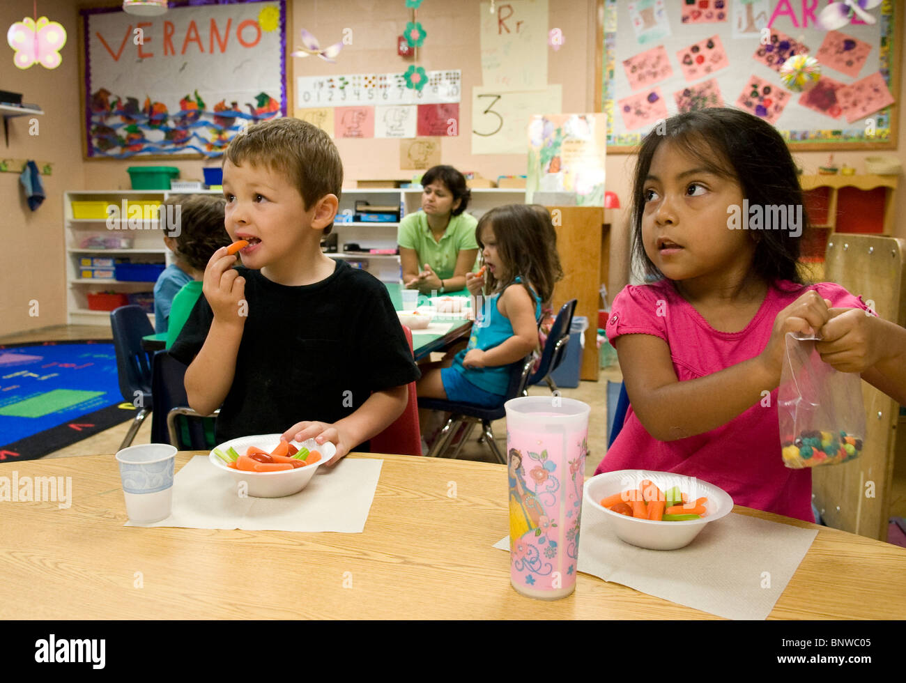 Hispanische Vorkindergarten-Alter Kinder essen Snacks in der Vorschule. Mädchen isst zuckerhaltige Getreide, während junge gesunde Karotten isst Stockfoto