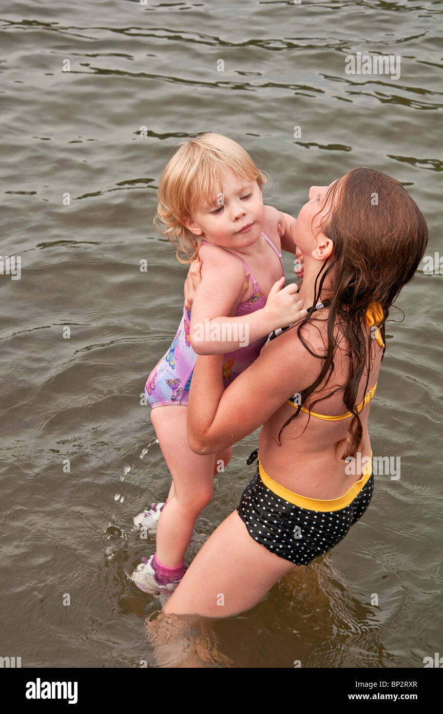 Kaukasisches 9 Jahres altes Mädchen ist heben und helfen eine jüngeren Kleinkind Mädchen im See sind in Badeanzügen Spaß. Stockfoto