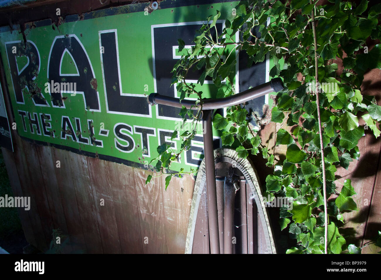 Ein Alter Ofen Emaille Werbeschild Raleigh die Ganzstahl-Fahrrad überwuchert von Efeu und unter beleuchtet Stockfoto