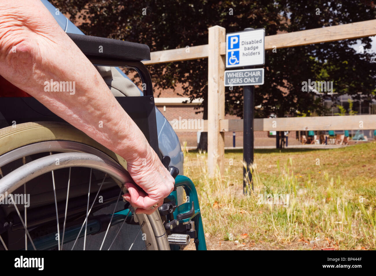 Ältere Frau im Rollstuhl durch ein Auto in behinderten Parkplatz durch ein Zeichen für blue badge Rollstuhlfahrer geparkt nur sitzen. England, Großbritannien, Großbritannien. Stockfoto