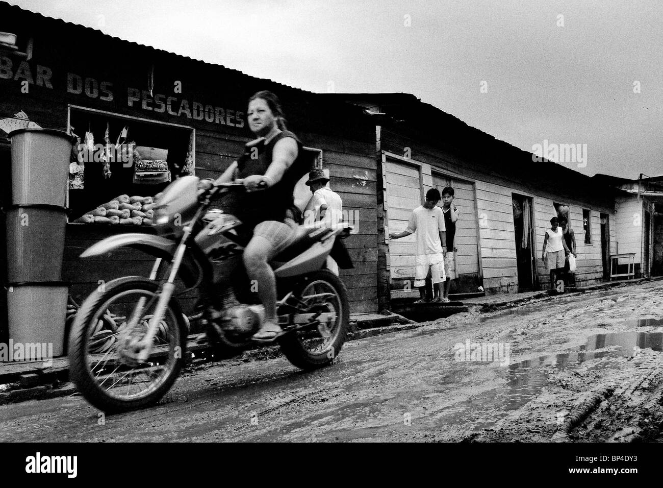 Eine Frau auf einem Motorrad durch die unbefestigten, schlammigen Straße in einer kleinen Fluss Gemeinde São Paulo de Olivença, Brasilien vorbei. Stockfoto