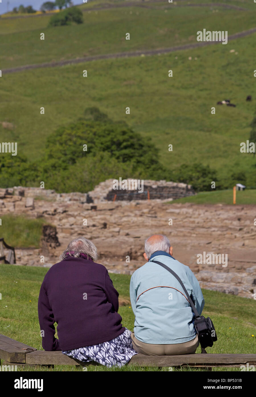 Ein älterer Mann und Frau sitzen auf einer Holzbank mit Blick auf die Ausgrabungen von Vindolanda, einer römischen Stadt in Northumbria Stockfoto