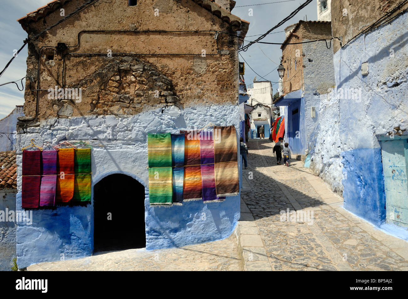 Straßenszene in Chefchaouen mit Anzeige der marokkanische Teppiche zum Verkauf hängen an den Wänden & blaue Häuser, Marokko Stockfoto