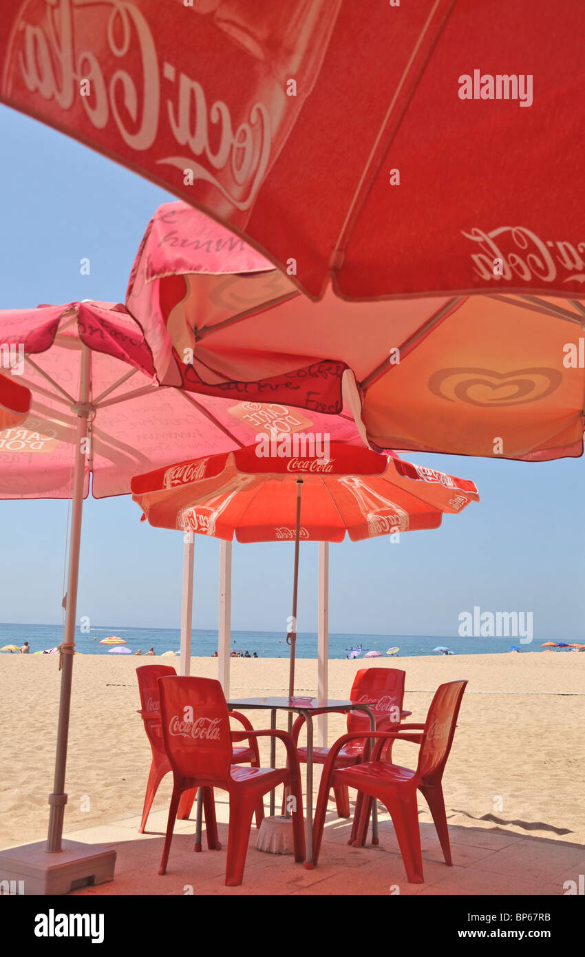 Rote Sonnenschirme Werbung für Coca Cola und Stühlen unter ihrem Schatten an einem Strand in Calella.Spain Stockfoto