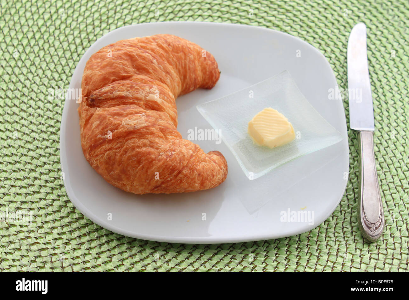 Goldene Croissant mit Orangenscheibe auf einem weißen Teller mit einem Messer und einem Stückchen Butter auf einer grünen Weide Tischset Stockfoto