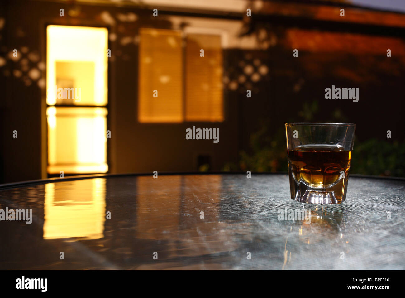 Bourbon Whiskey in Schnapsglas auf Glastisch mit Haus im Hintergrund in der Nacht Stockfoto