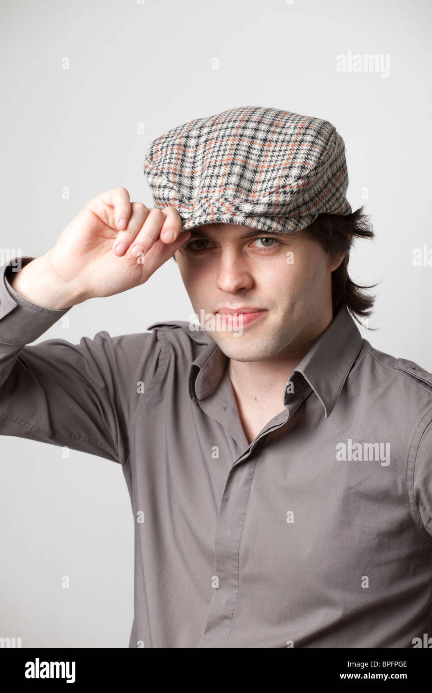 Junger Mann in einem grauen Hemd und karierter flacher Mütze, kippt seinen Hut und blickt selbstbewusst in die Kamera in einer Studioaufnahme. Stockfoto