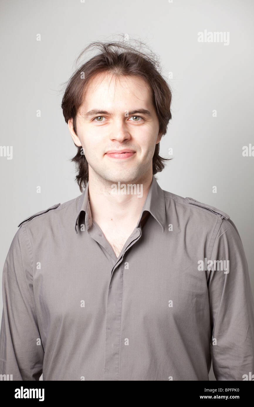Junger Mann mit braunen Haaren, der ein graues Hemd trägt und mit einem leichten Lächeln direkt in die Kamera blickt. Stockfoto