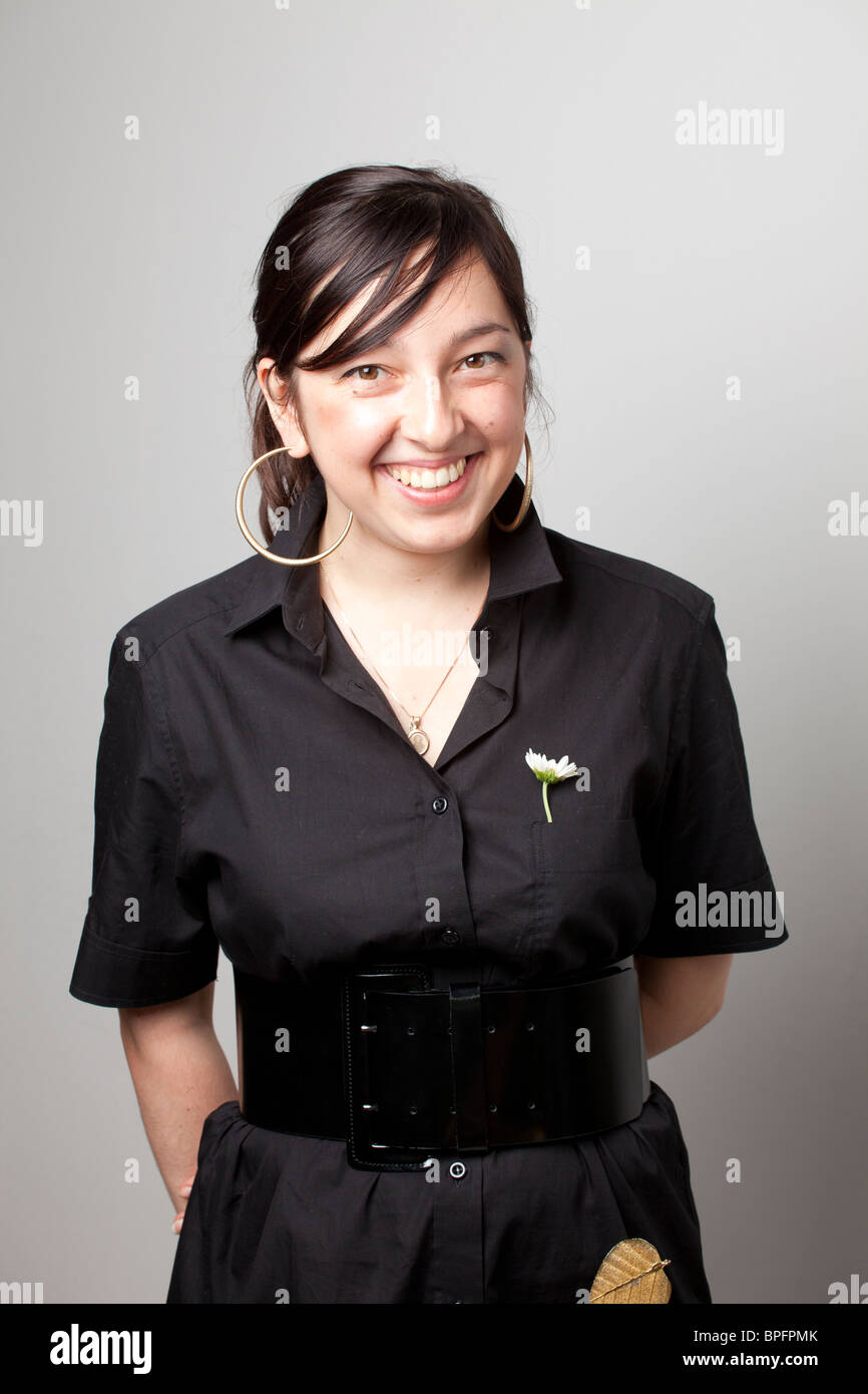 Eine junge Frau in einem schwarzen Kleid mit einer weißen Blume in der Tasche, die hell vor einem neutralen Hintergrund lächelt. Stockfoto
