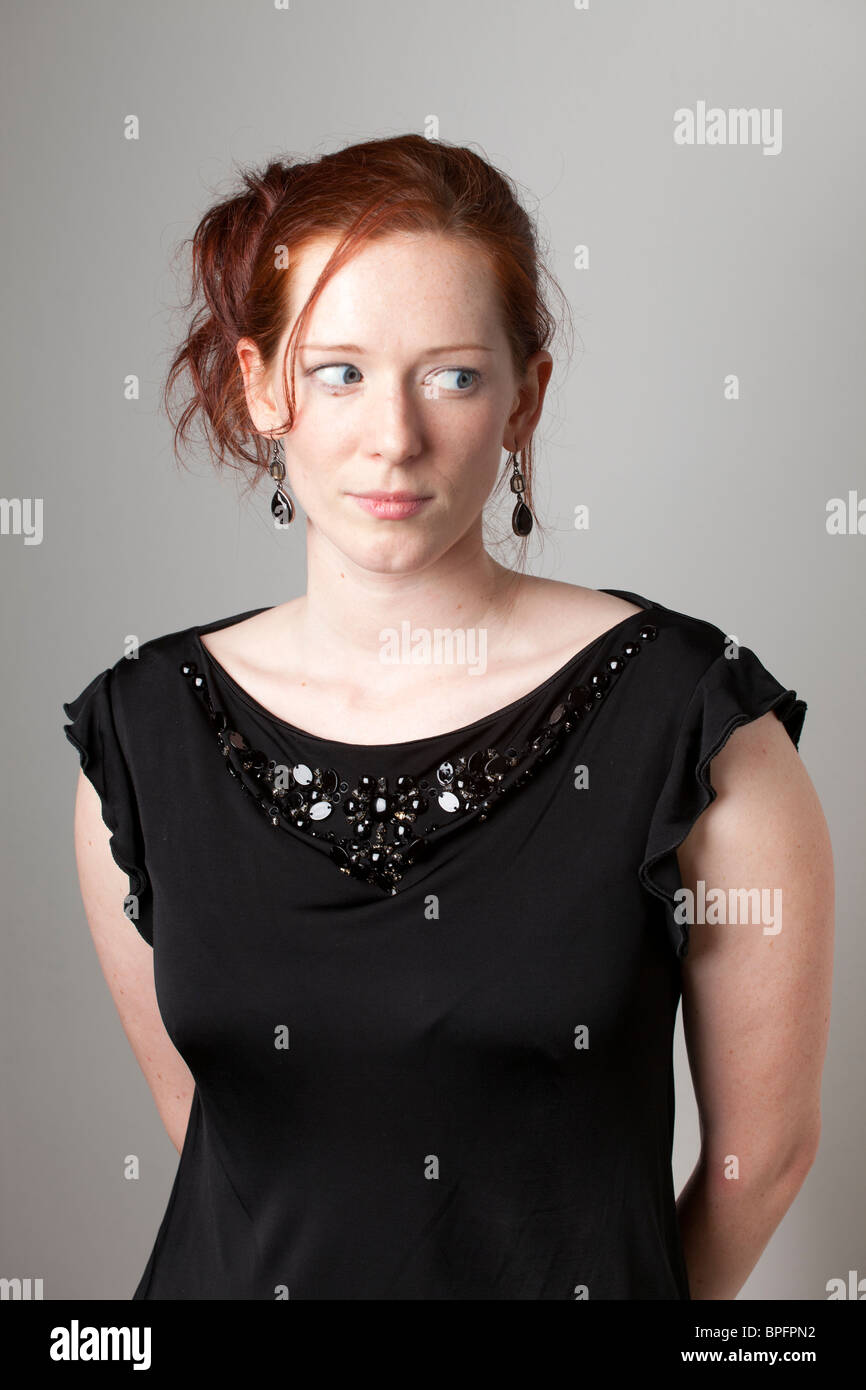 Junge Frau in einem schwarzen Oberteil mit verziertem Ausschnitt, der seitlich vor einem neutralen Hintergrund schaut. Stockfoto