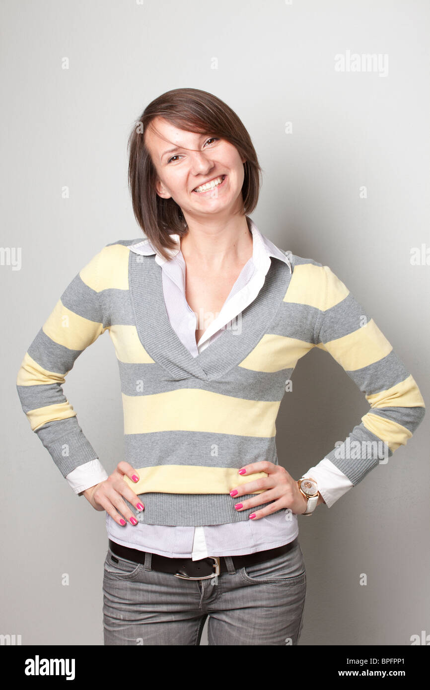Junge Frau in einem gestreiften Pullover und weißem Hemd, die selbstbewusst mit Händen auf den Hüften vor neutralem Hintergrund lächelt. Stockfoto