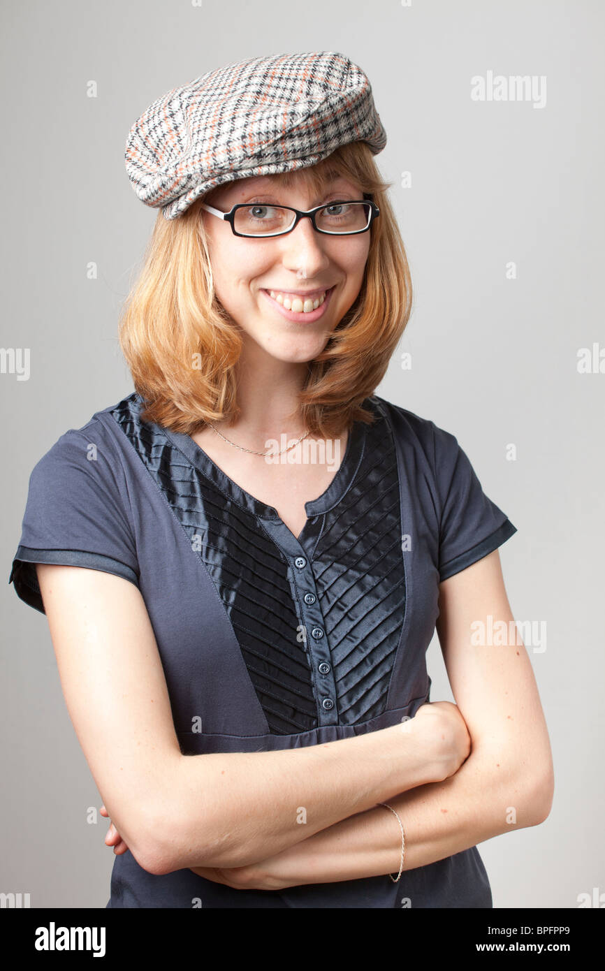 Junge Frau in blauem Hemd und karierter flacher Mütze, mit Brille und einem Lächeln mit überkreuzten Armen vor neutralem Hintergrund. Stockfoto