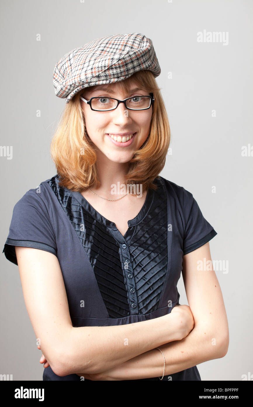 Junge Frau in blauem Hemd und karierter flacher Mütze, mit Brille und einem Lächeln mit überkreuzten Armen vor neutralem Hintergrund. Stockfoto