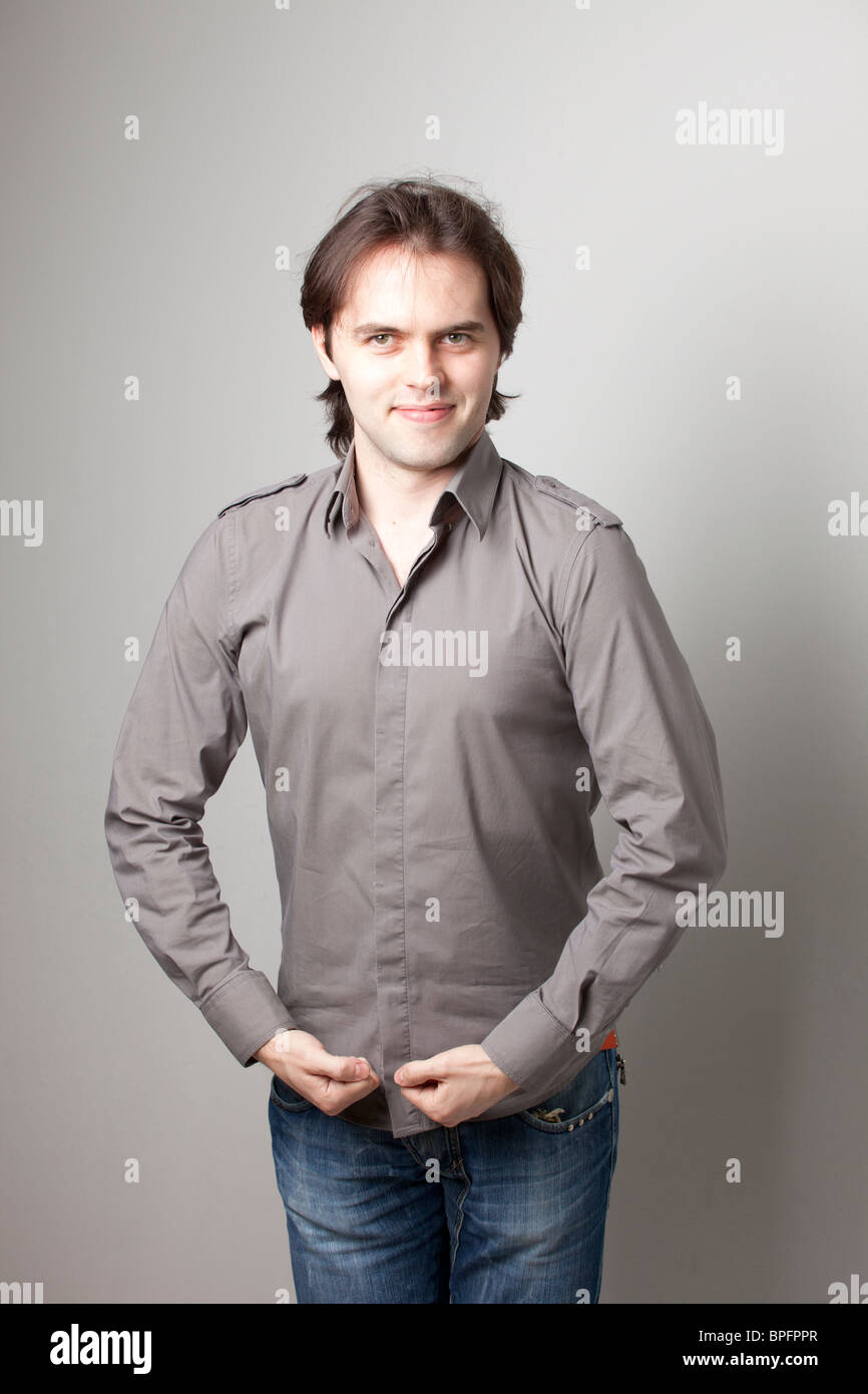 Junger Mann in grauem Hemd und Jeans, der selbstbewusst vor neutralem Hintergrund lächelt. Stockfoto