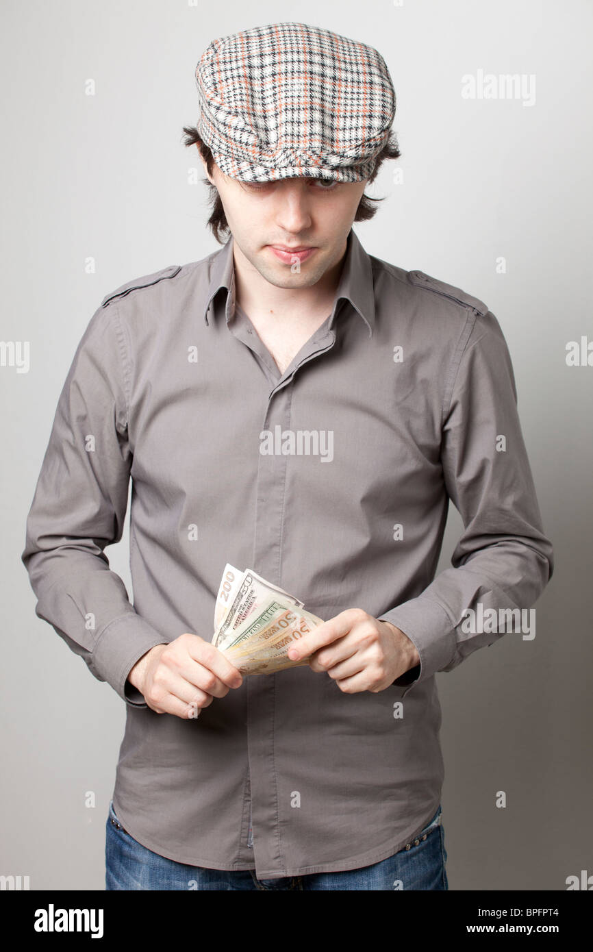 Junger Mann in einem grauen Hemd und karierter flacher Mütze, der auf Geld in seinen Händen vor einem neutralen Hintergrund herabblickt. Stockfoto