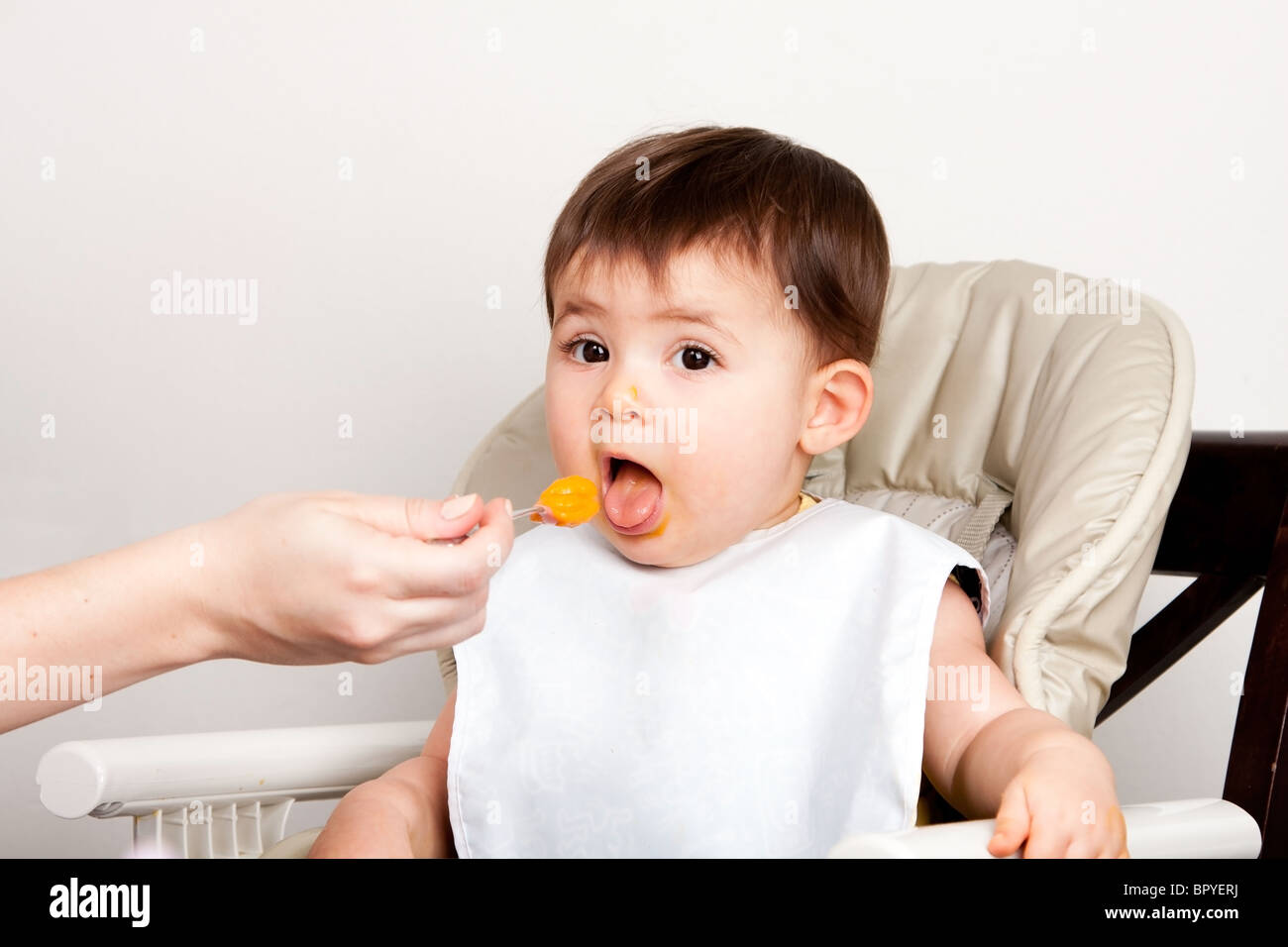 Schönes glückliches Baby Säugling Junge Mädchen mit Mund öffnen chaotisch Orangen-Püree vom Löffel zu essen. Stockfoto