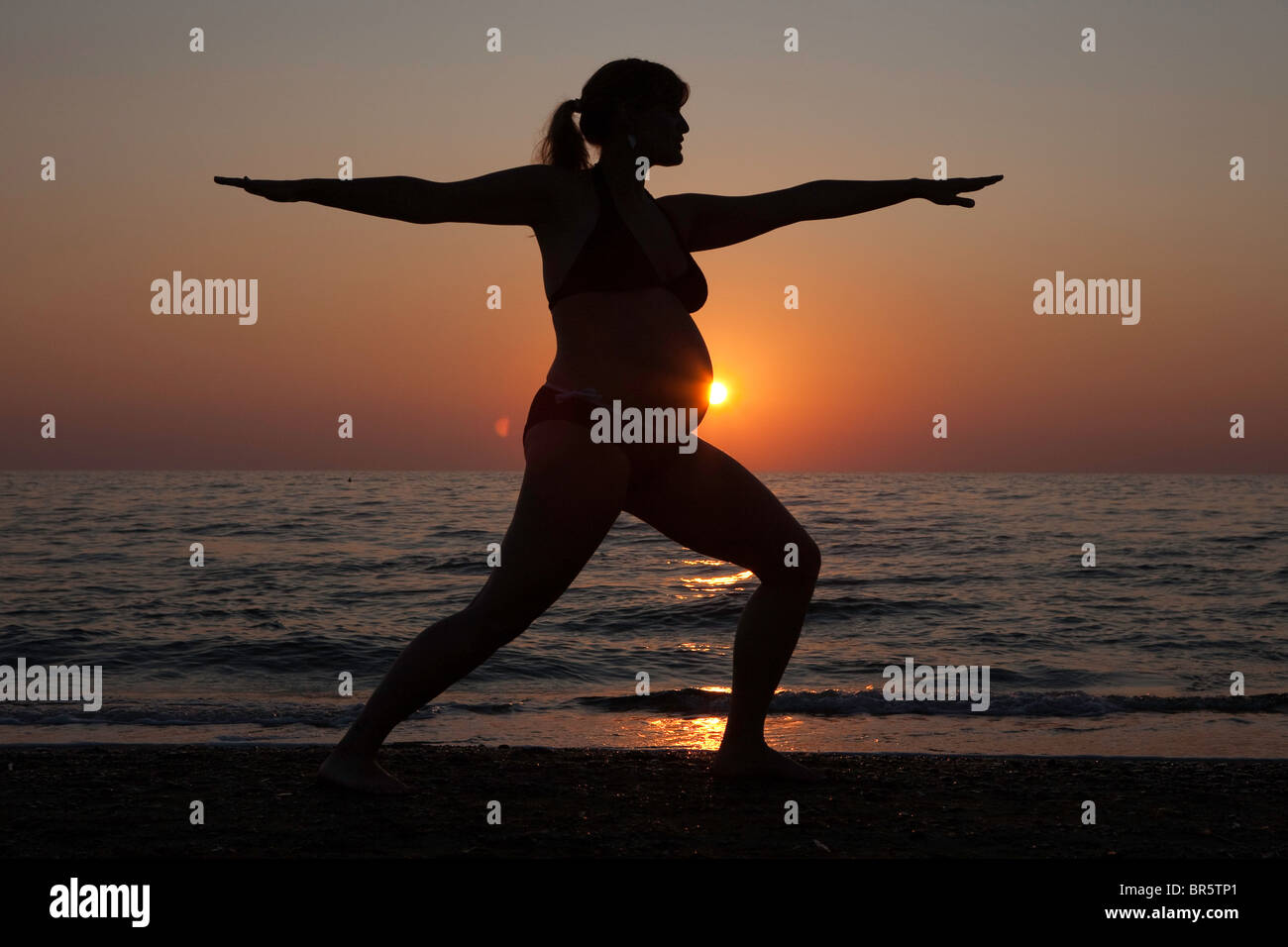 Eine schwangere Frau praktiziert Yoga am Strand in der Toskana, Silhoutted von der untergehenden Sonne. Stockfoto
