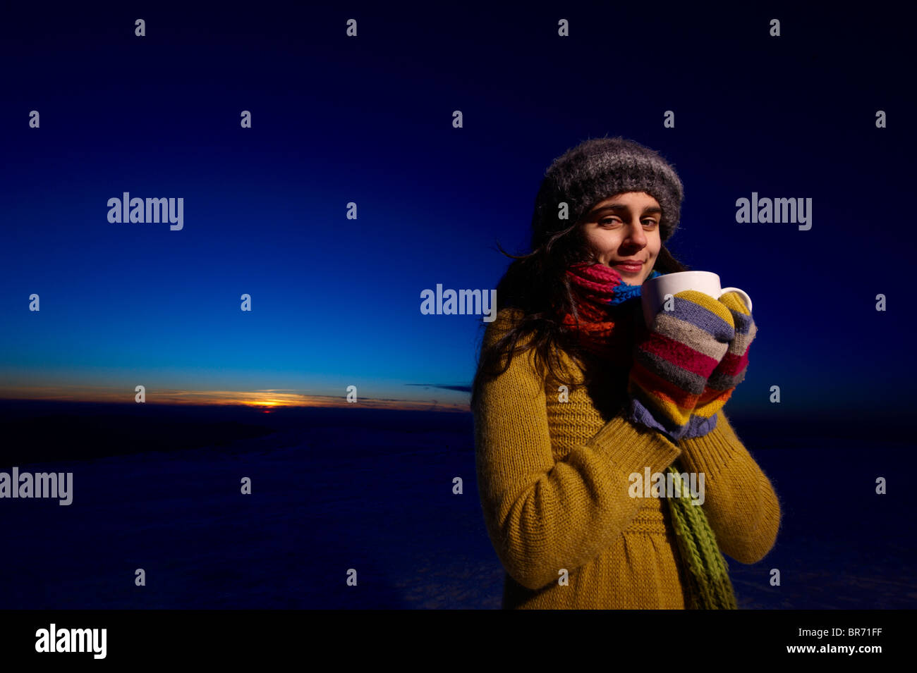 Eine lächelnde junge Frau hält eine Tasse heiße Schokolade vor einem Sonnenuntergang Himmel Stockfoto