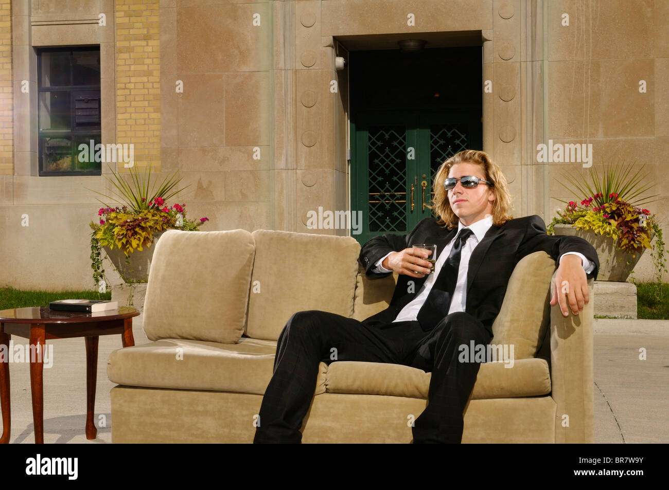 Reichen jungen Mann mit langen blonden Haaren im Anzug sitzen auf einer Couch mit einem Drink auf der Terrasse in einem Herrenhaus Stockfoto