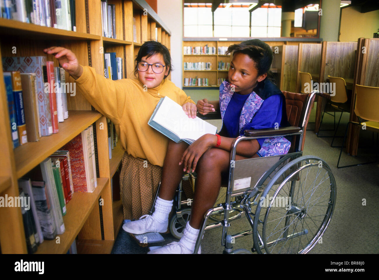 Hispanischen Mädchen hilft schwarzes Mädchen Handicap Rollstuhl Schule Bibliothek Hilfe Freund helfen Stockfoto