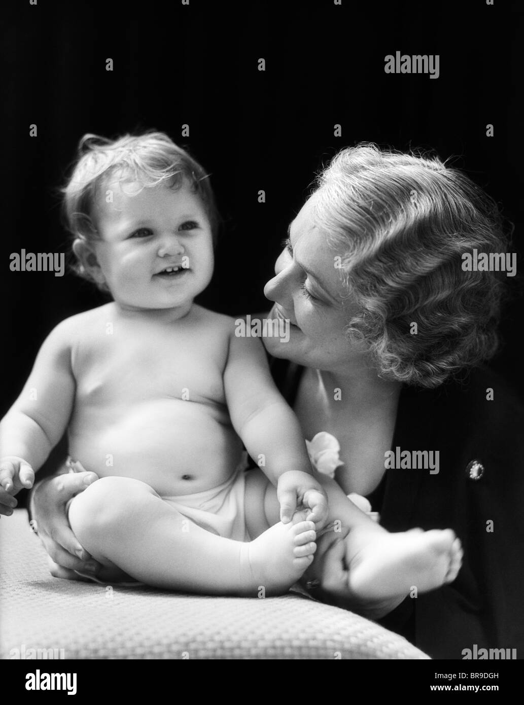 1930ER JAHREN BLONDE FRAU MUTTER LÄCHELND BETRACHTET MAN BABY AUF KISSEN SITZEND Stockfoto