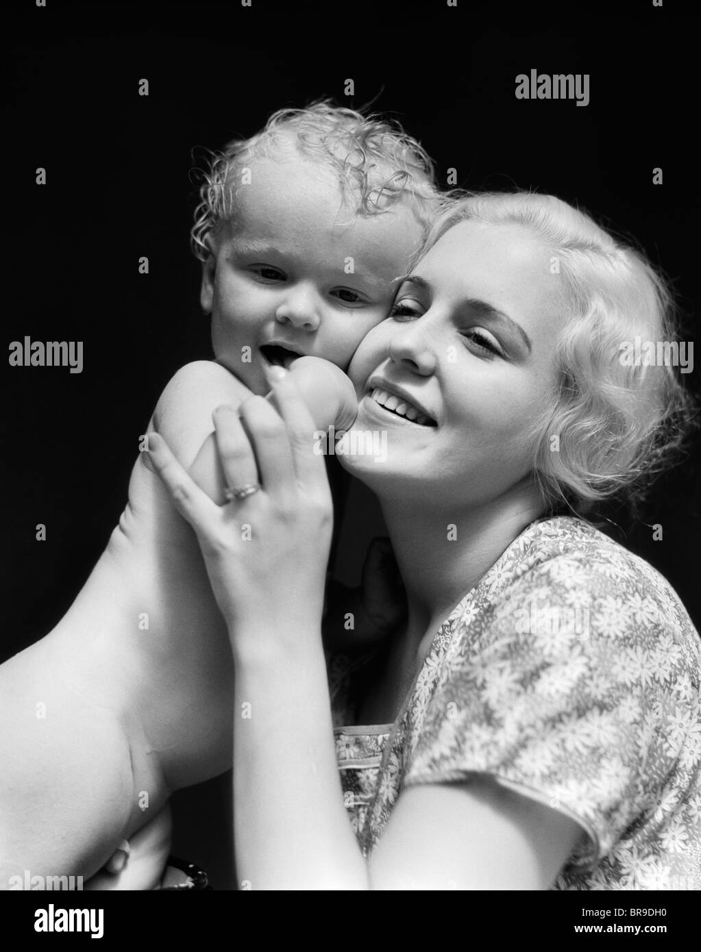 1930ER JAHREN BLONDE FRAU MUTTER LÄCHELND HOLDING BABY AN WANGE Stockfoto