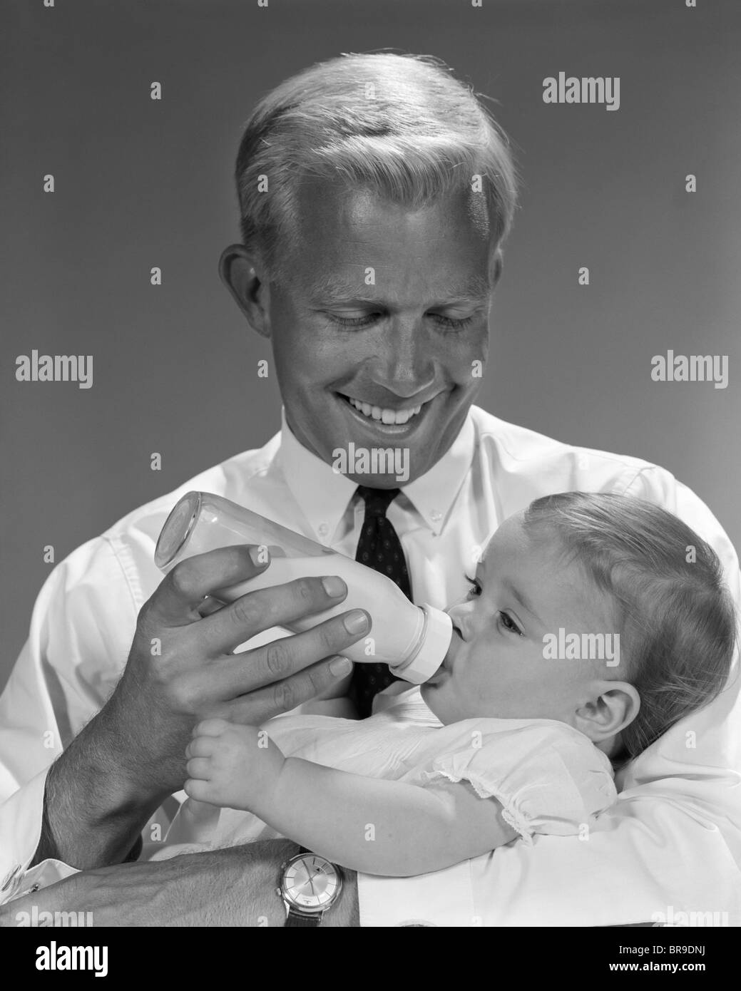 1960ER JAHREN LÄCHELND BLOND VATER KIND MILCH IN GLASFLASCHE BABY FÜTTERN Stockfoto