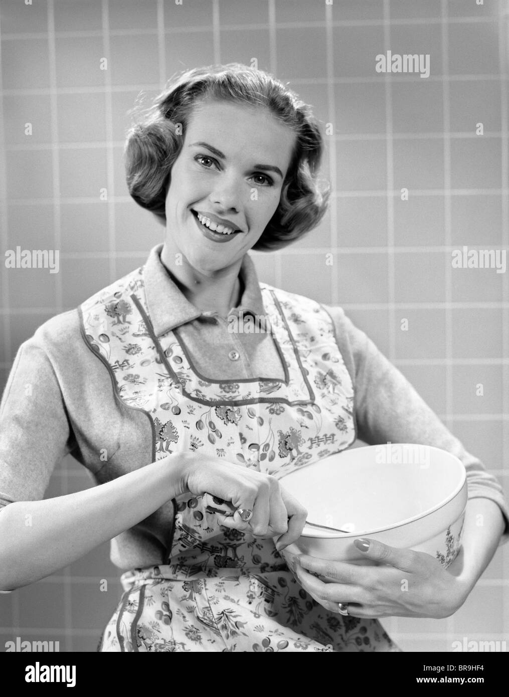 1950ER JAHREN LÄCHELNDE FRAU TRÄGT SCHÜRZE IN KÜCHE HOLDING MISCHENDE SCHÜSSEL UND LÖFFEL BLICK IN DIE KAMERA Stockfoto