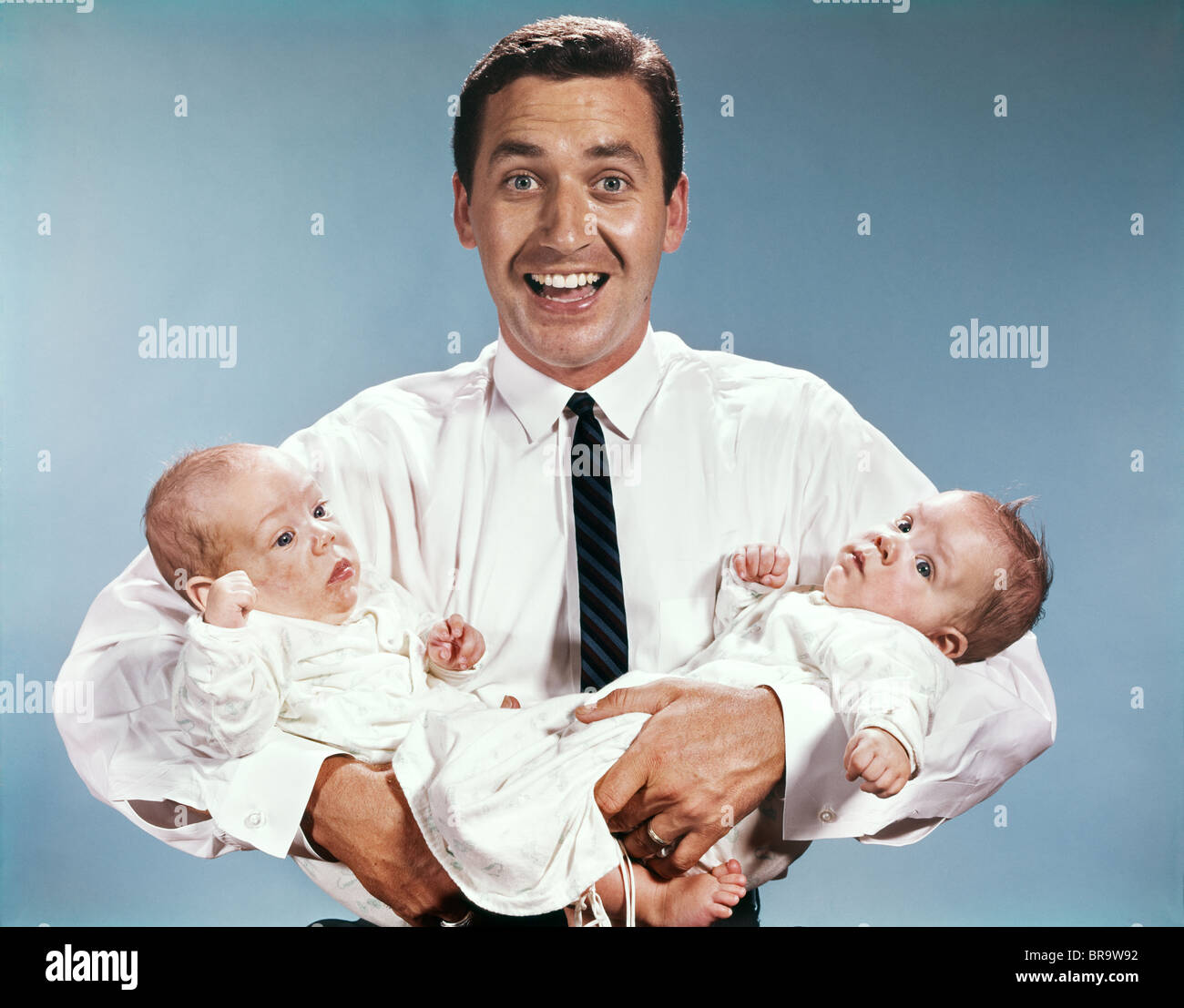 1960ER JAHREN LÄCHELND MANN VATER BLICK AUF KAMERA HOLDING TWIN BABYS KLEINKINDER Stockfoto
