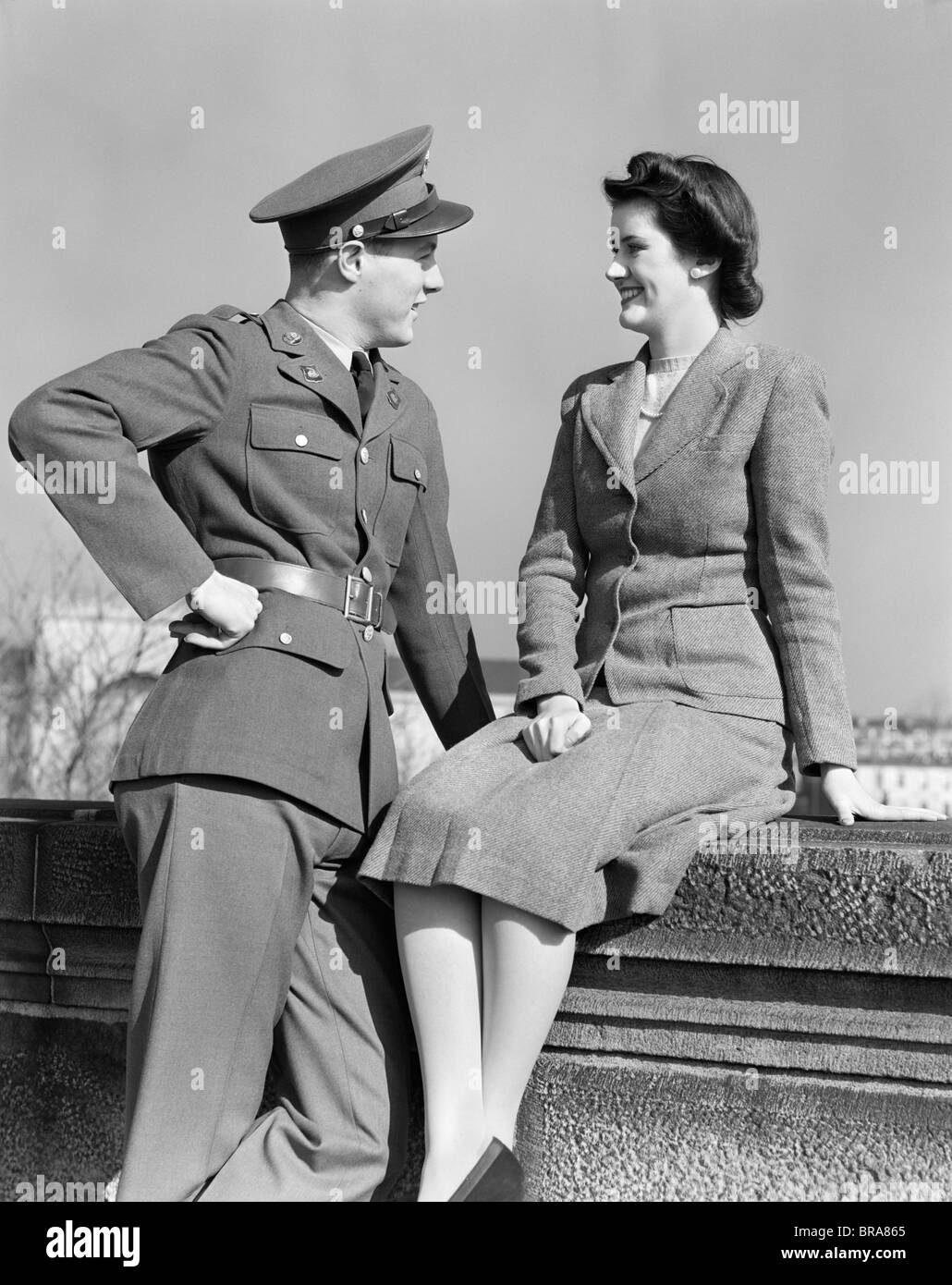 1940ER JAHREN GLÜCKLICHE PAAR MANN TRAGEN ARMEE UNIFORM FRAU SITZEN AUF STEINMAUER Stockfoto