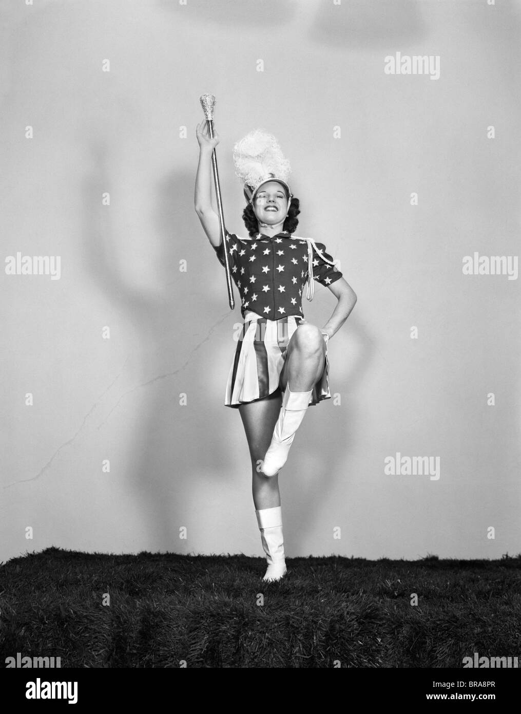 1940ER JAHRE MAJORETTE POSIERT MIT SCHLAGSTOCK MIT BLICK NACH VORNE TRAGEN PATRIOTISCHEN STARS AND STRIPES EINHEITLICHE MIT WEIßEN STIEFELN UND GEFIEDERTE HUT Stockfoto