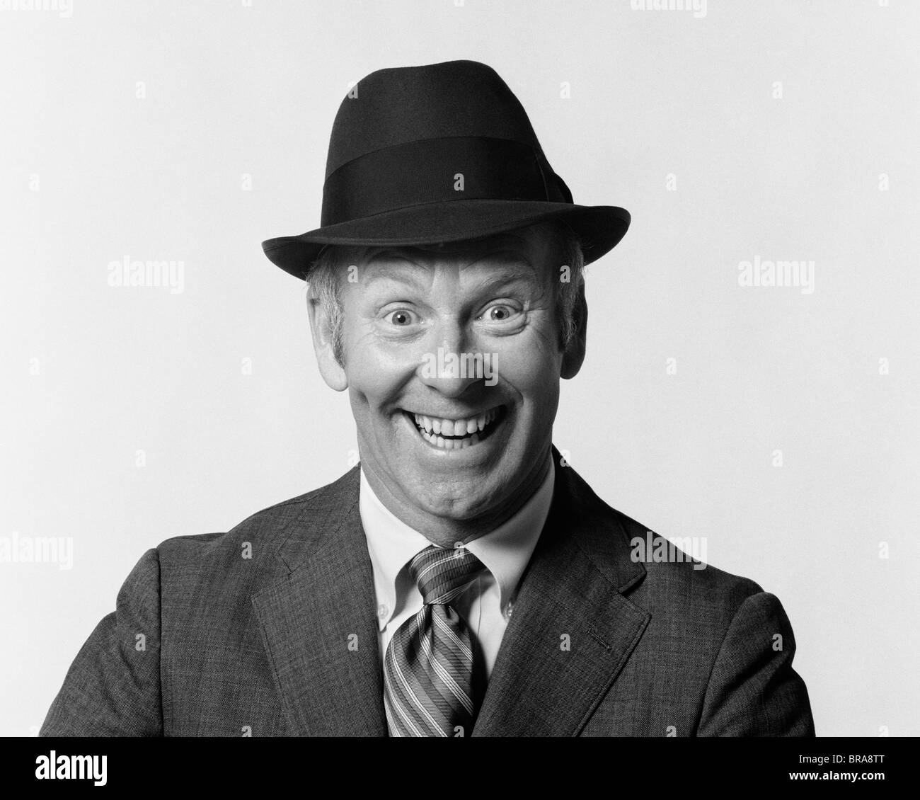 1960ER JAHRE MANN LÄCHELND TRAGEN ANZUG UND HUT MIT DUMMEN EIFRIG GESICHTSAUSDRUCK Stockfoto